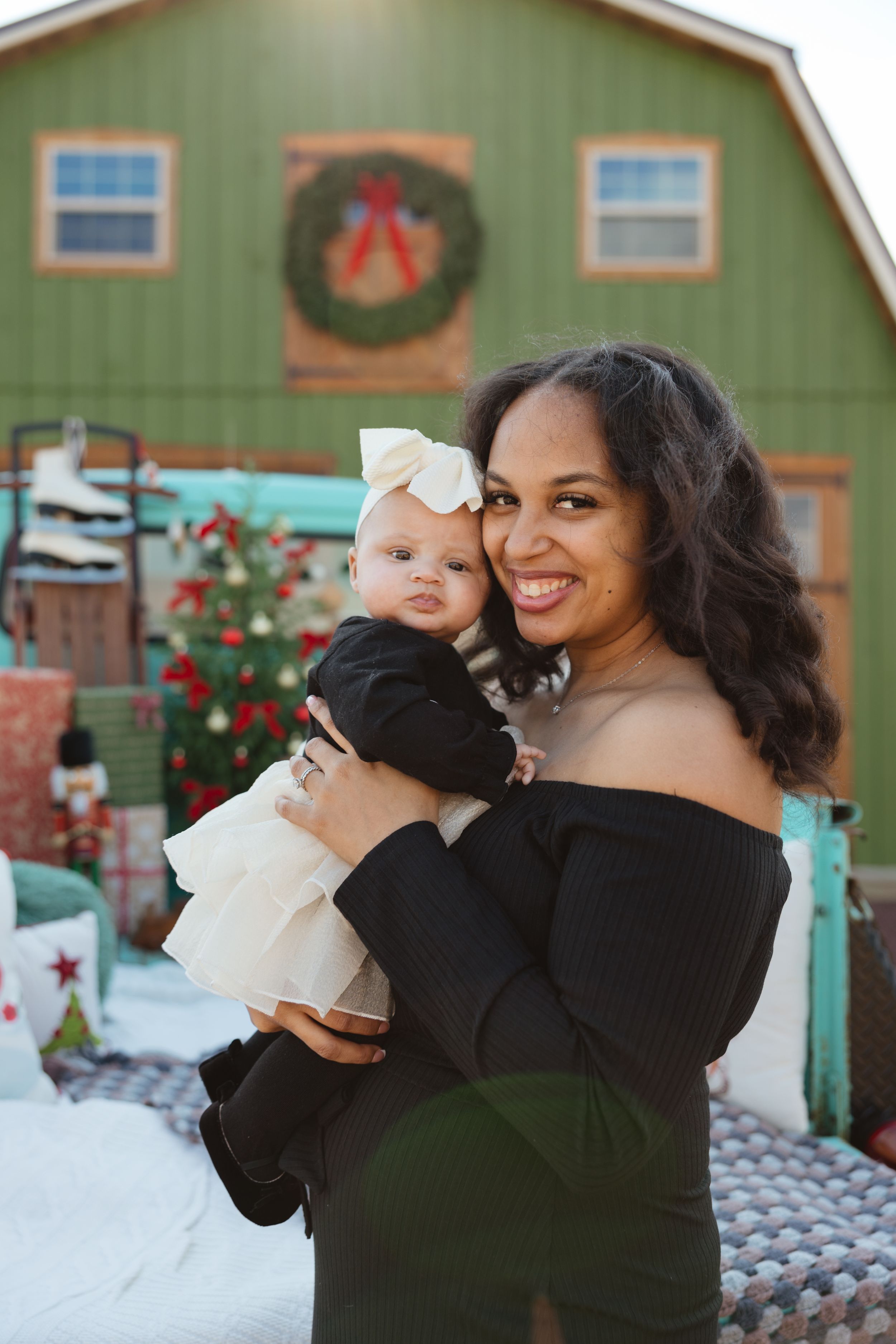 A smiling woman holds a baby in a white bow outdoors near a festive green barn with a Christmas wreath.