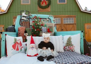 Baby in black outfit sits in vintage teal truck bed with Santa pillows and Christmas decorations outside a green barn.
