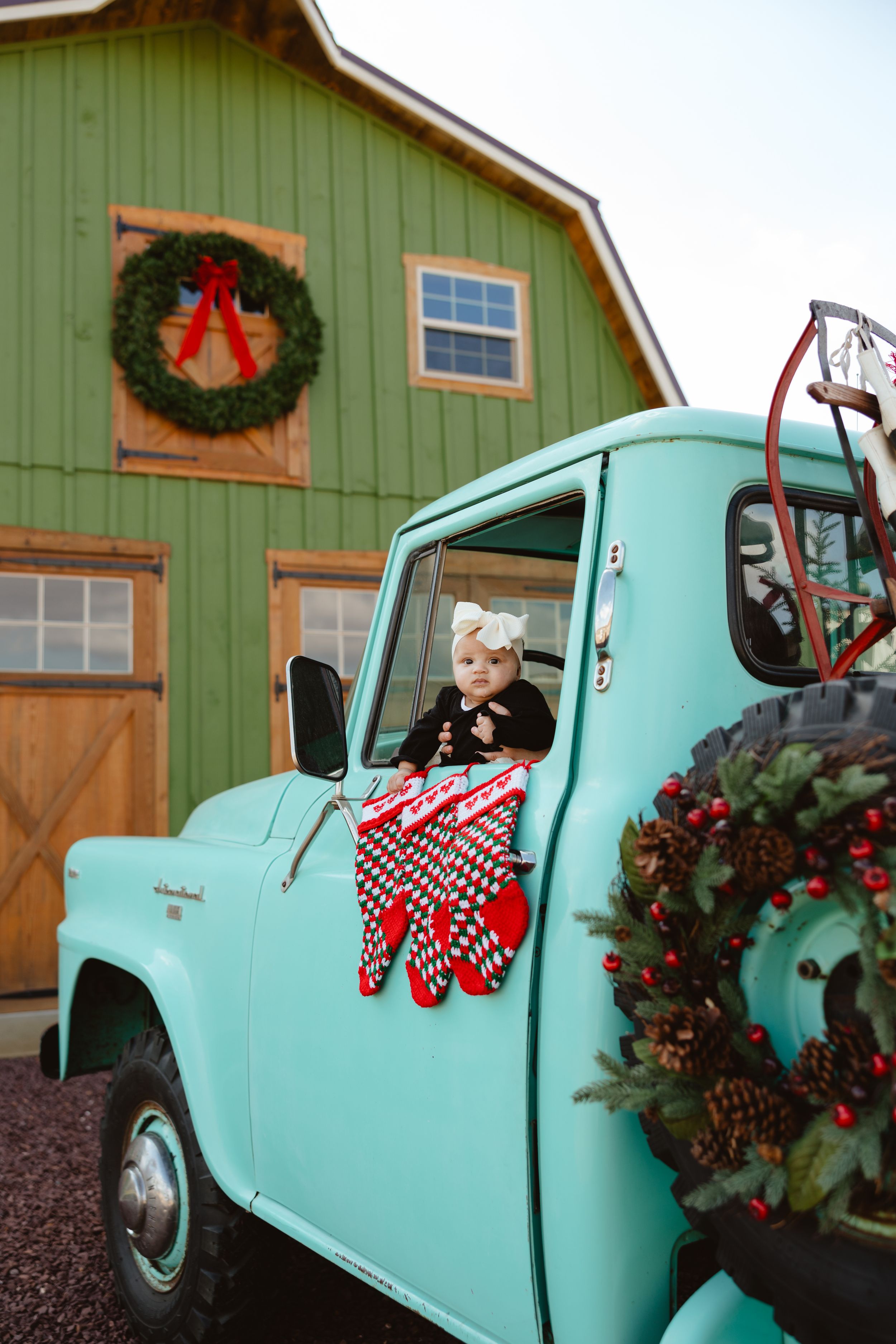 Baby in teal vintage truck decorated with Christmas wreaths and stockings in front of a green barn.