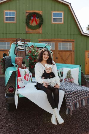 Woman holding baby sits on vintage teal truck bed decorated for Christmas outside green barn.