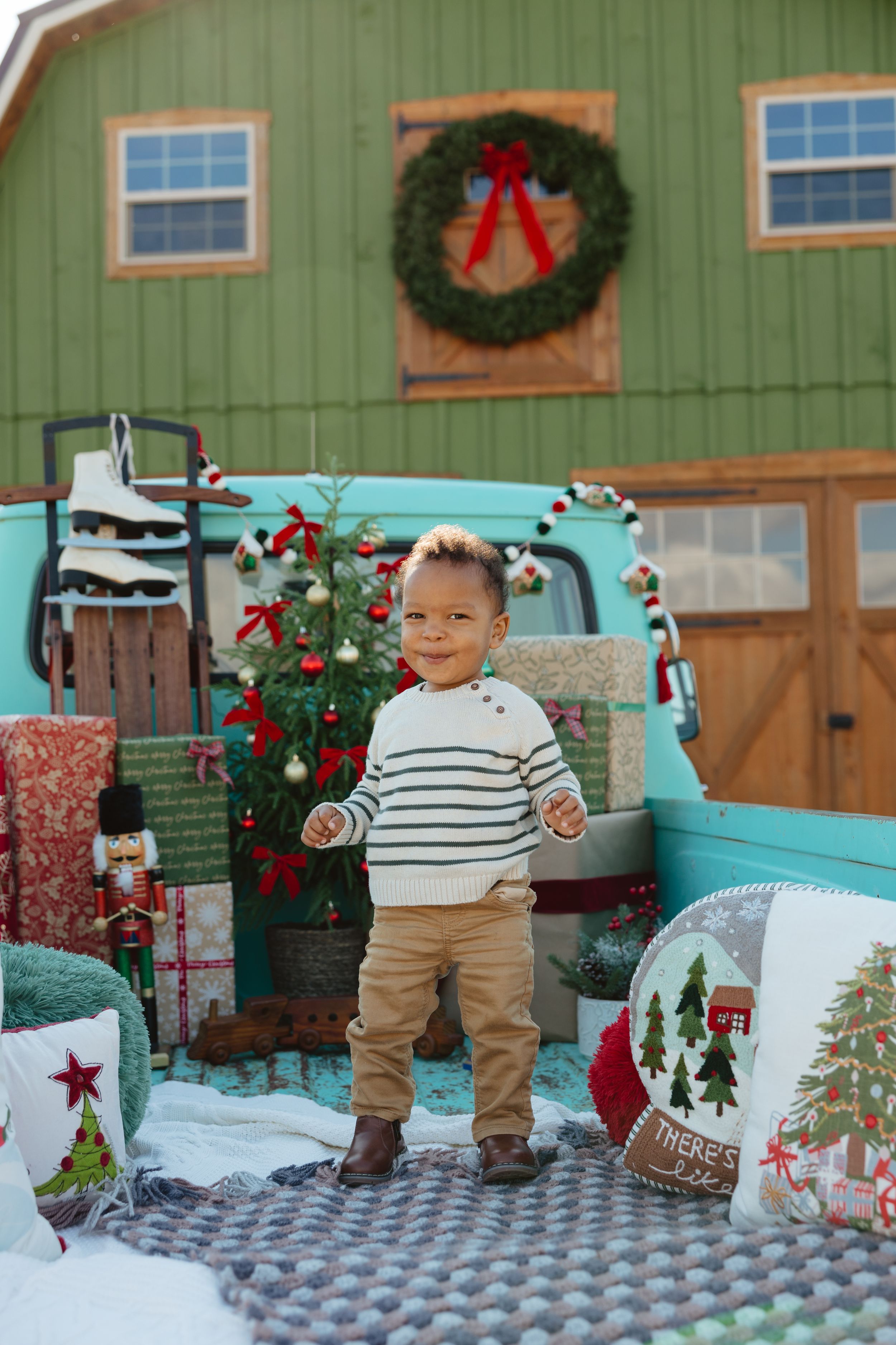 Toddler boy in striped sweater poses before a festive teal truck and green barn adorned with Christmas wreath and decorations.
