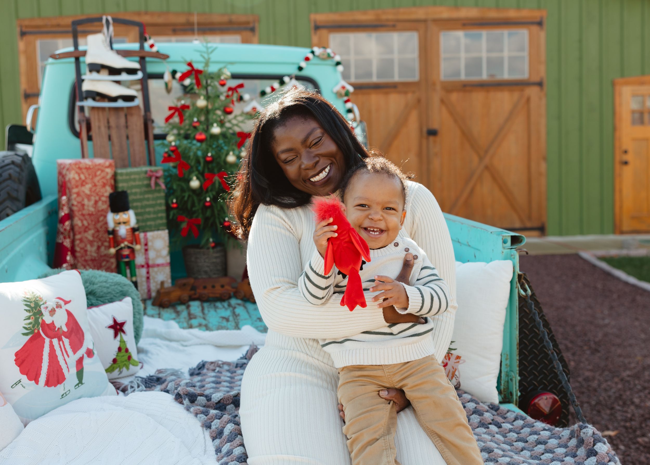 A joyful mother holds her laughing baby boy while opening a red Christmas gift near a decorated tree and teal truck.