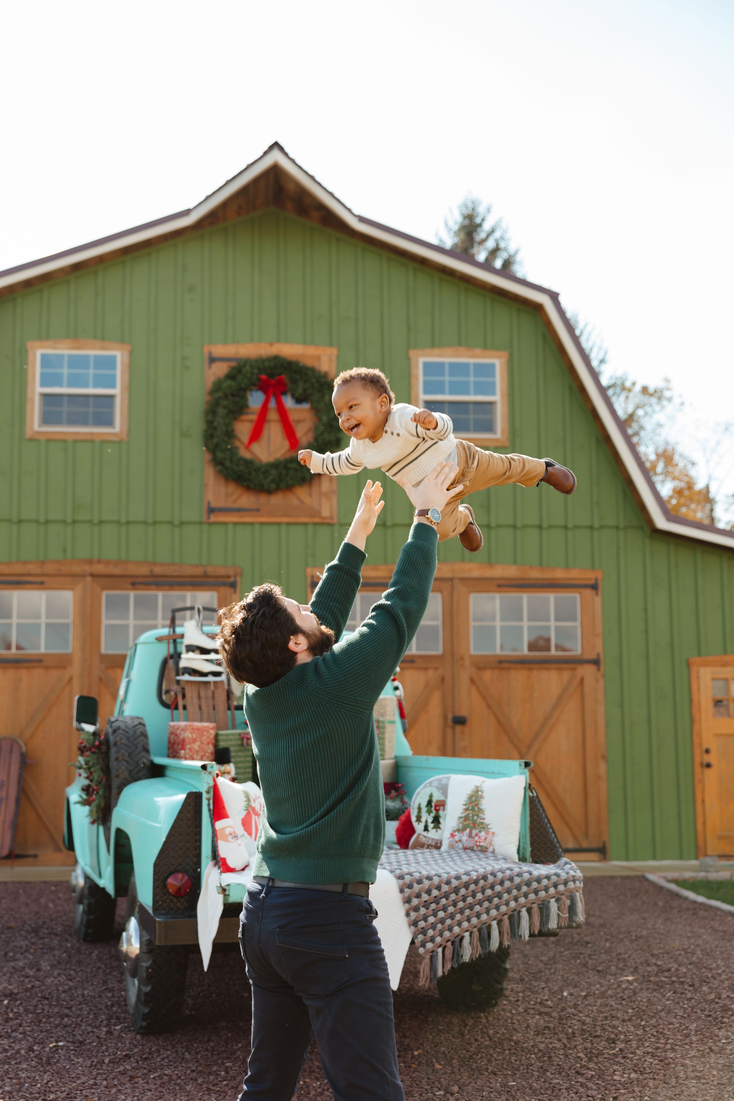 Father joyfully lifting baby in front of green Christmas barn with wreath and vintage teal truck.