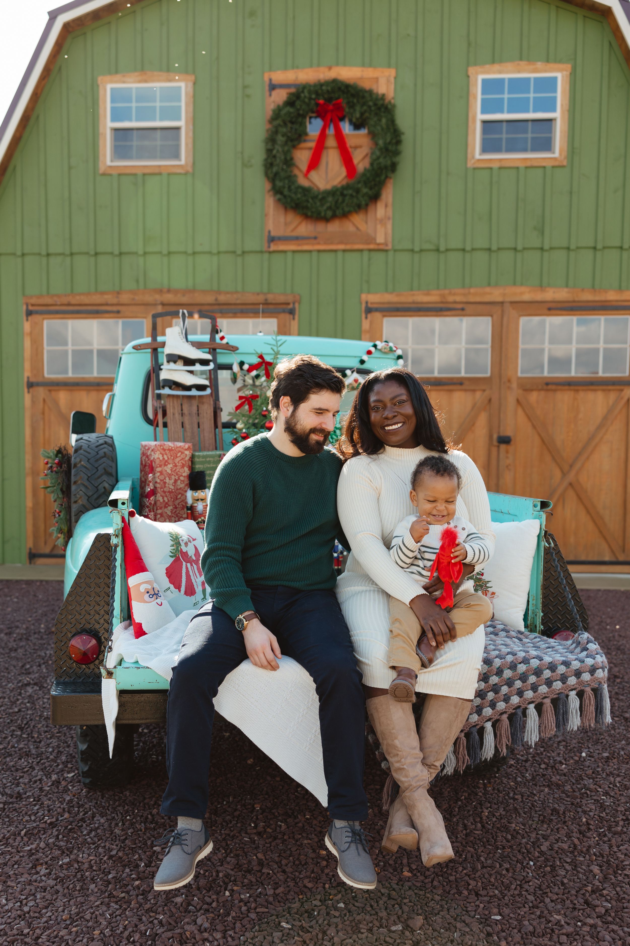 Interracial family with baby sitting on vintage teal truck bed before green barn with Christmas wreath decoration.