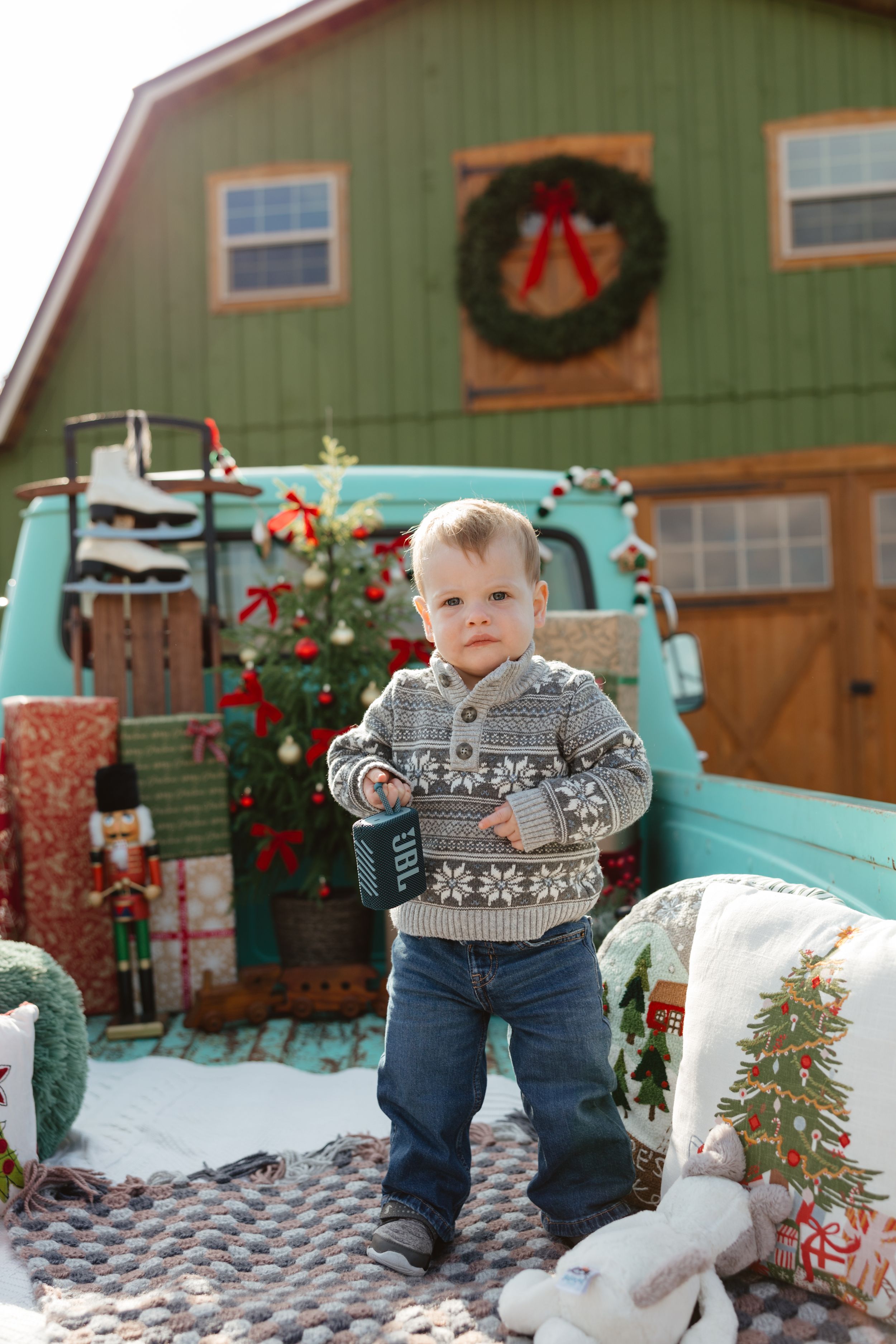 Toddler boy in Nordic sweater and jeans stands in truck bed with Christmas barn backdrop and holiday decorations.