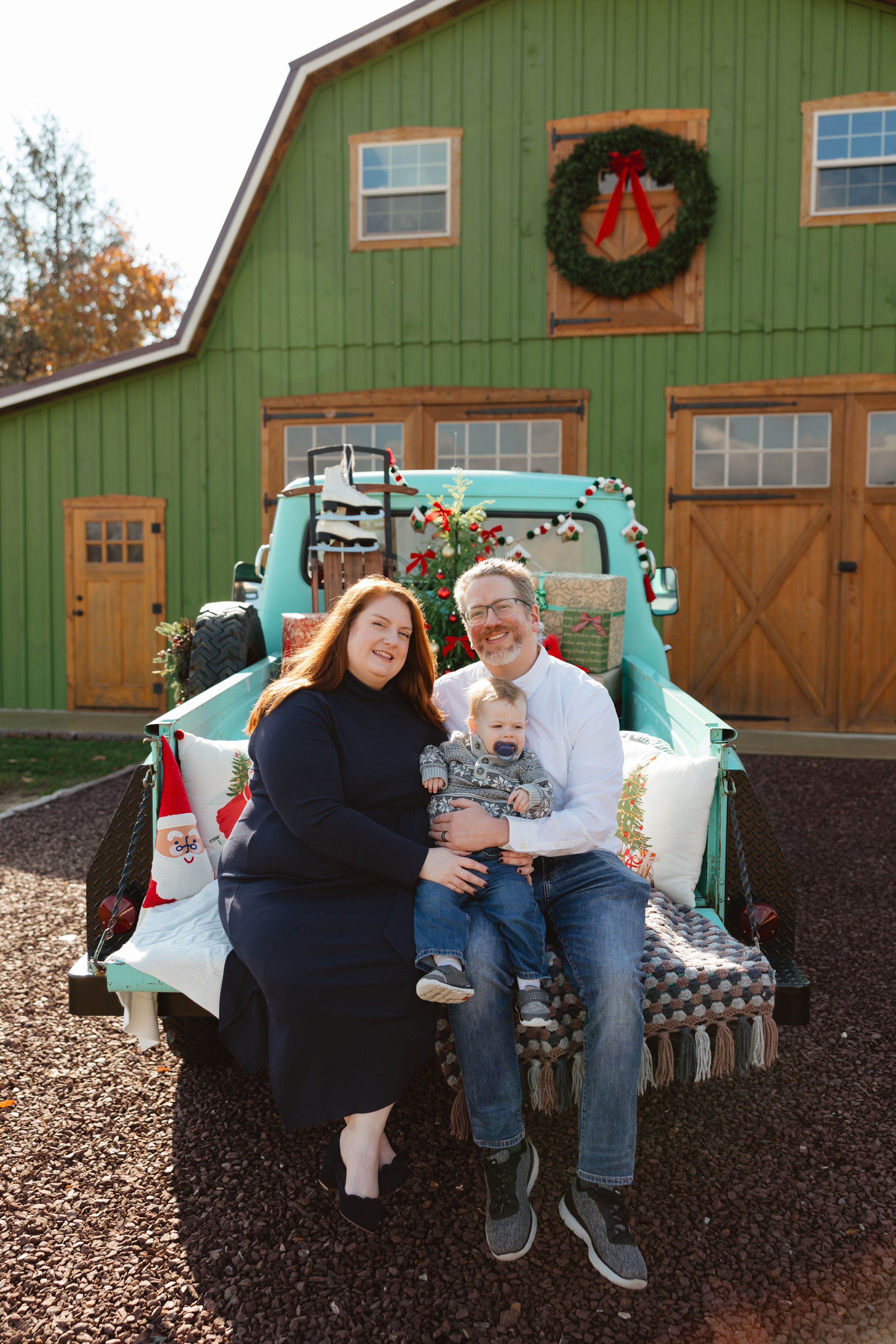 Family of three sitting in vintage teal truck bed before green barn with Christmas wreath holiday decor.