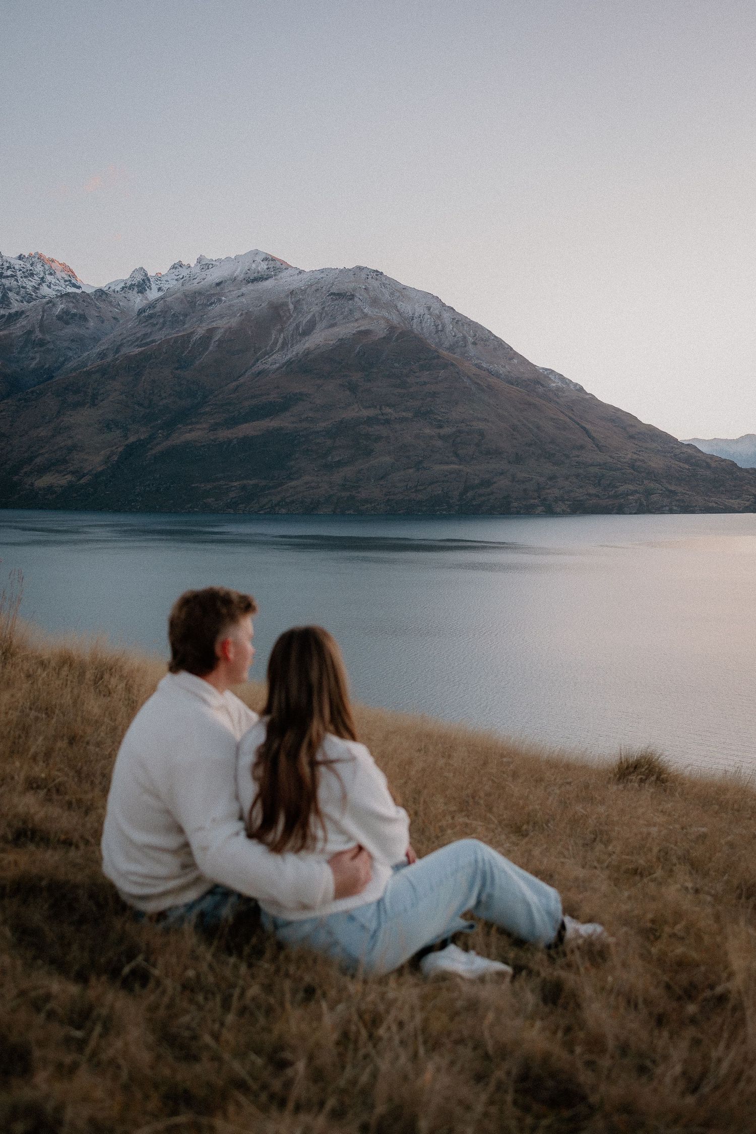 Couple sitting on grassy hillside embracing, overlooking serene mountain lake with snow-capped peaks at dusk.