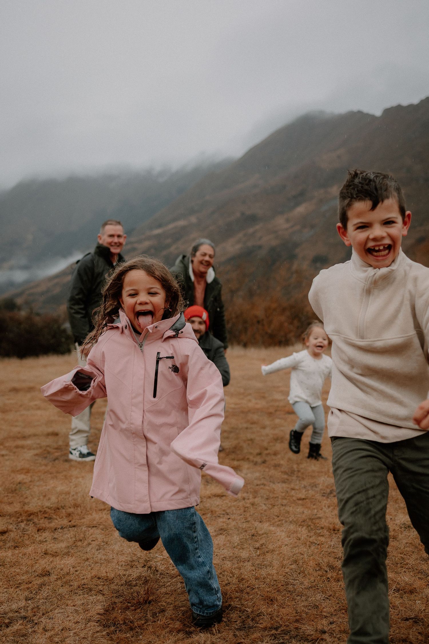 Two children laugh and run through a mountain meadow while their family follows behind on a cloudy day.