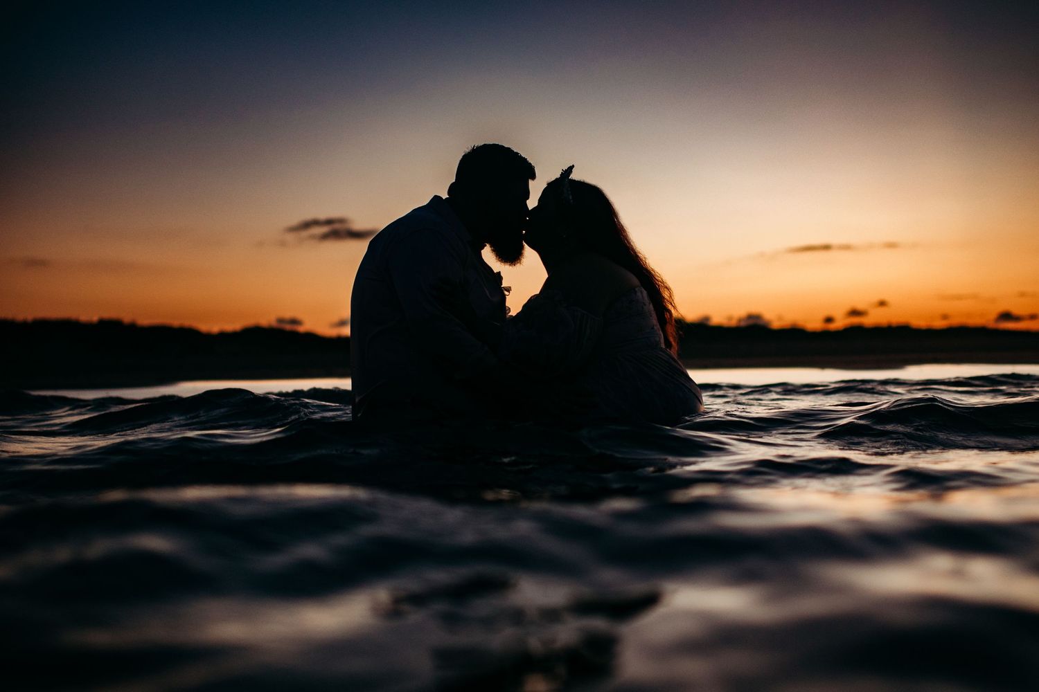 Romantic silhouette of couple kissing in ocean water during stunning golden sunset.