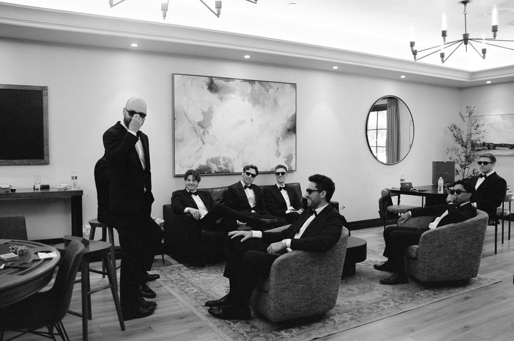 Groom and groomsmen in tuxedos relaxing in elegant lounge, black and white wedding photo.