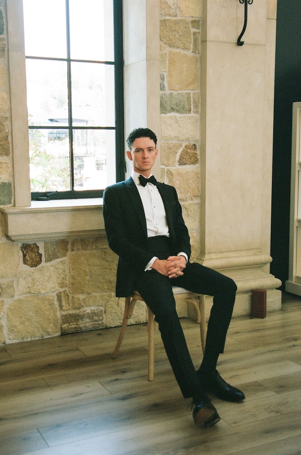 Groom in black tuxedo with bow tie seated by stone wall window in elegant venue.