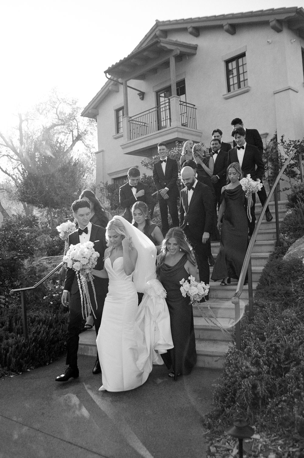 Wedding party descends outdoor stairs at elegant venue in black and white photo.