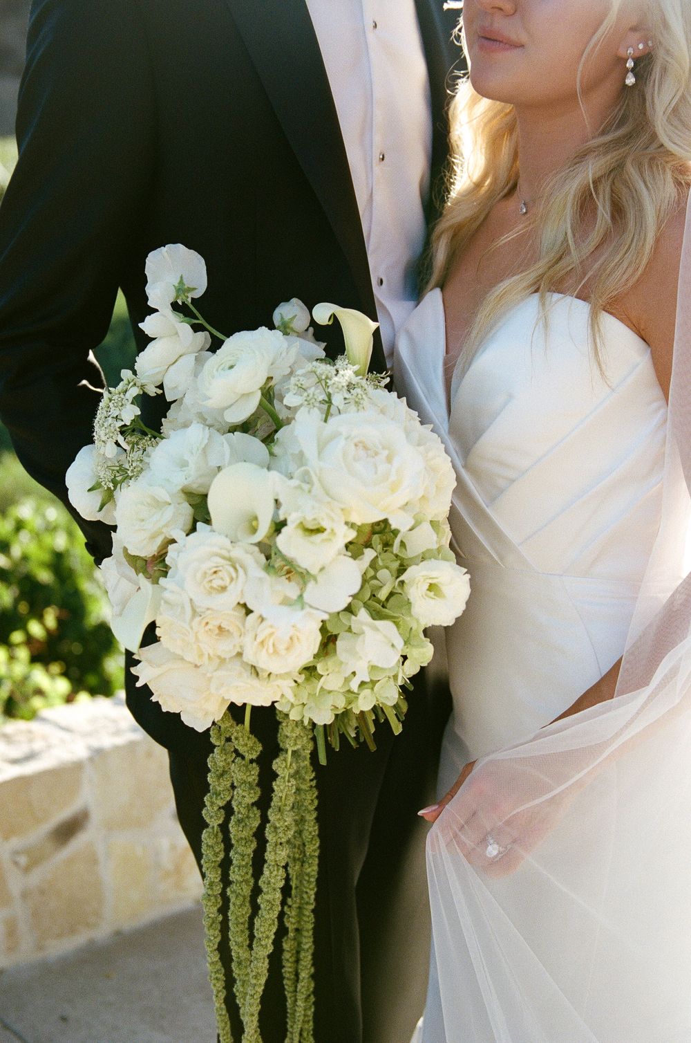 Bride in white strapless gown holds lush white rose and hydrangea bouquet with green amaranthus beside suited groom.