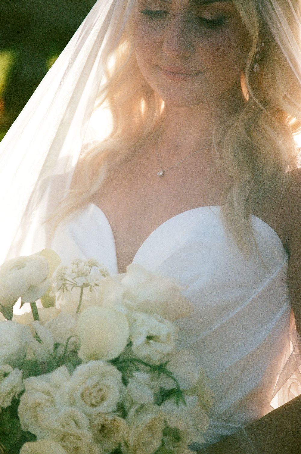 Blonde bride in strapless gown holds white rose bouquet under flowing veil.