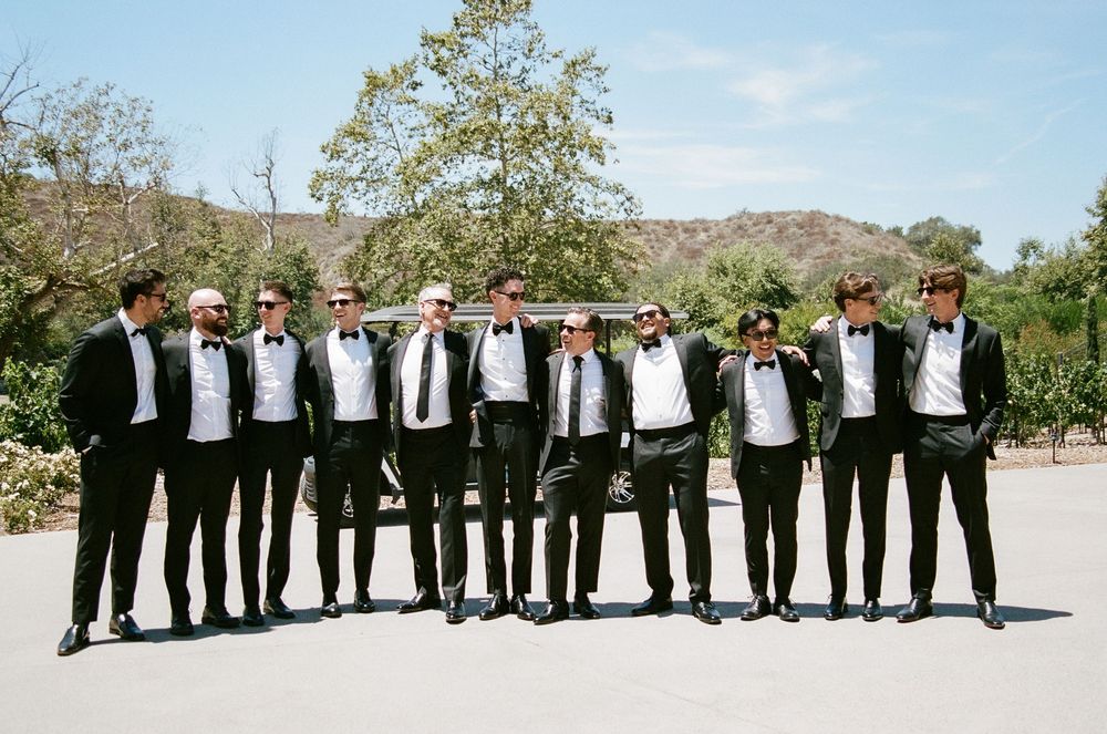 Groomsmen in matching black tuxedos and bow ties posing outdoors in a row on a sunny wedding day.