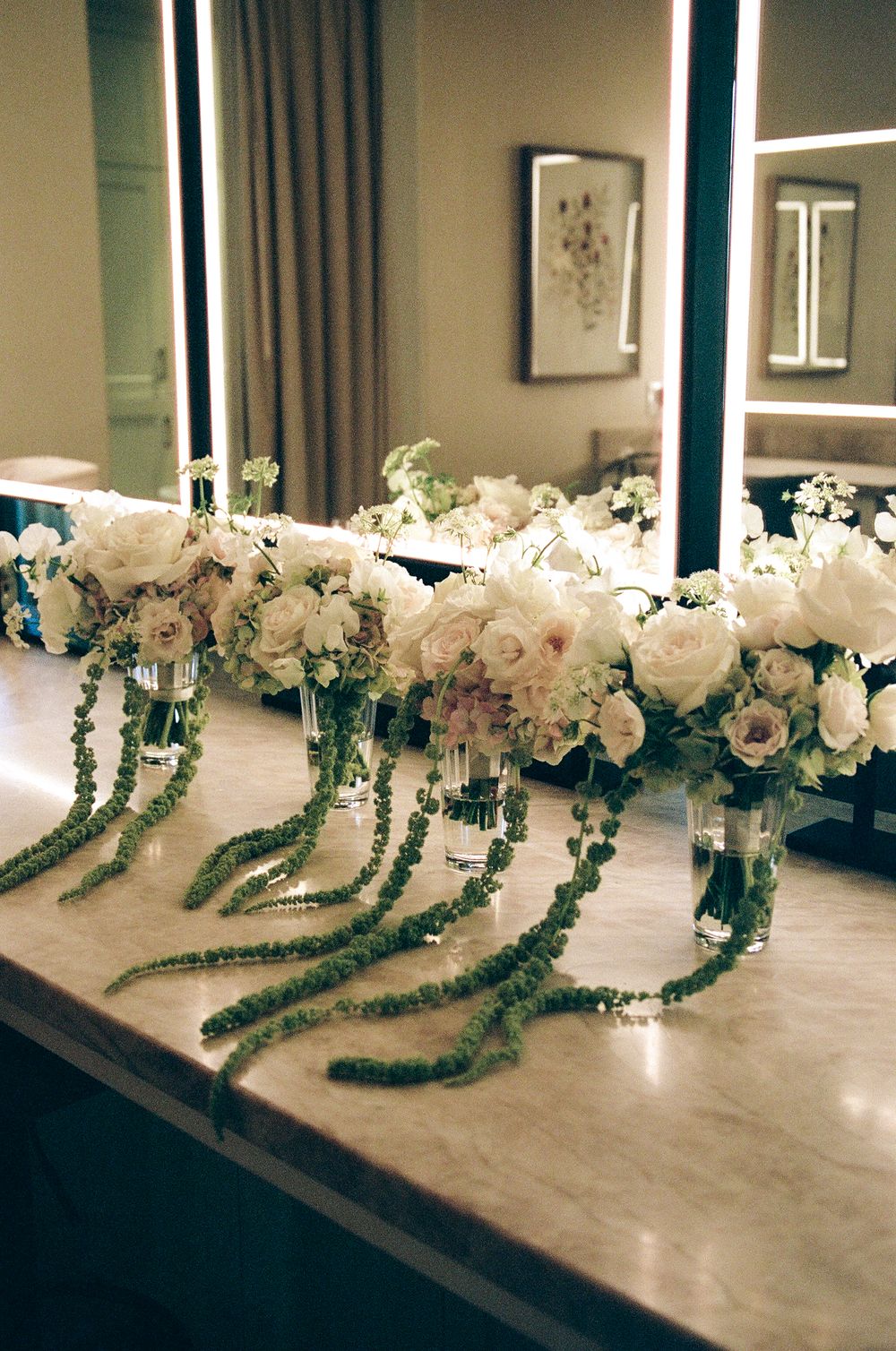 Elegant white floral arrangements with cascading green amaranthus in glass vases displayed on a marble counter.