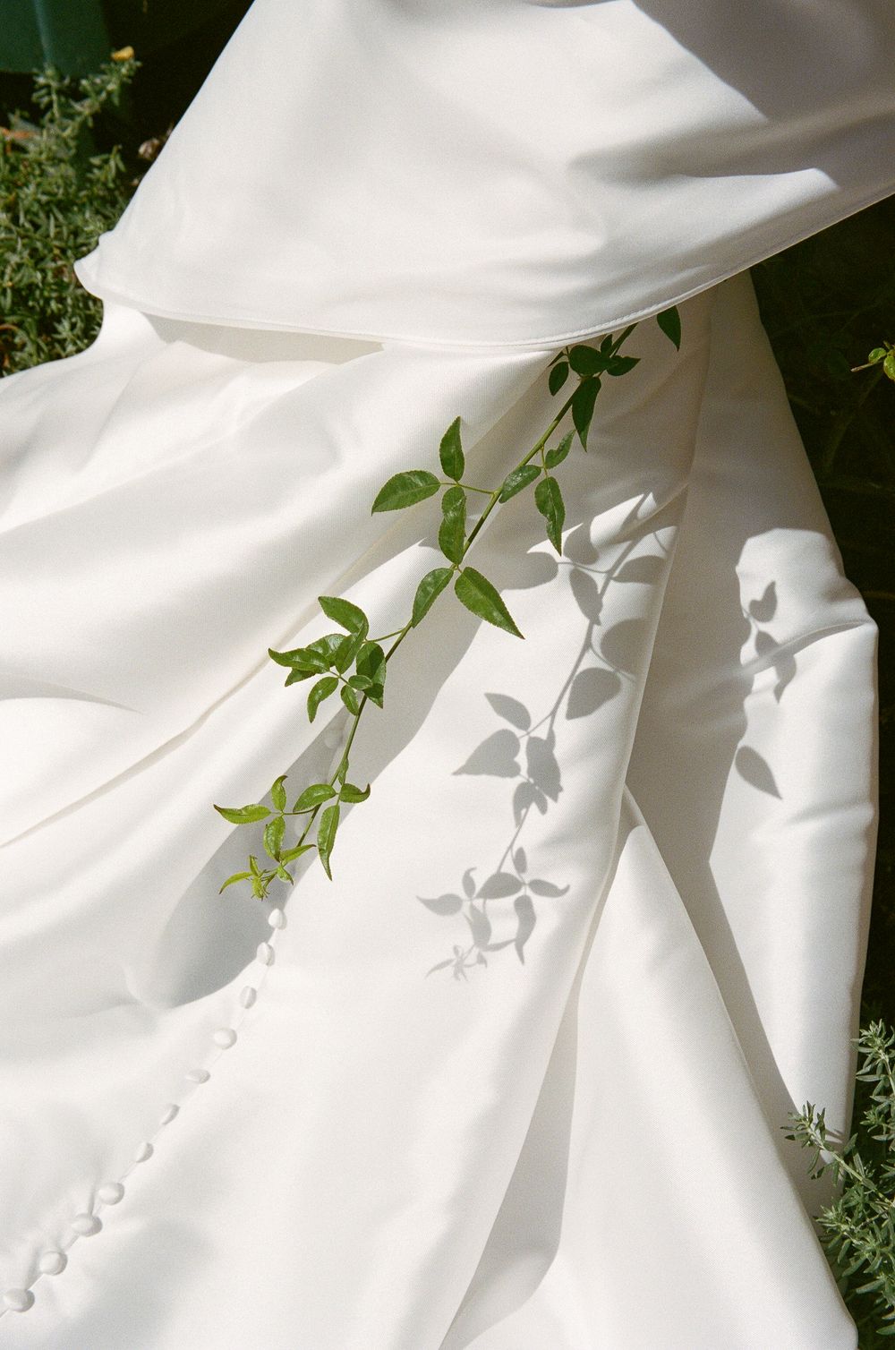 Elegant white wedding dress train laid on green grass with a delicate leafy branch casting shadows.
