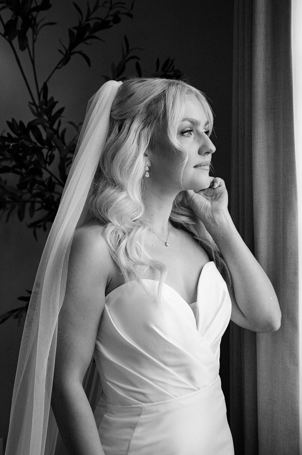 Elegant bride in strapless gown and veil gazes out window in black and white portrait photo.