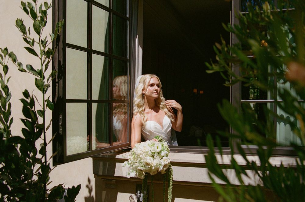Elegant bride in strapless gown holds white bouquet by French doors outdoors.