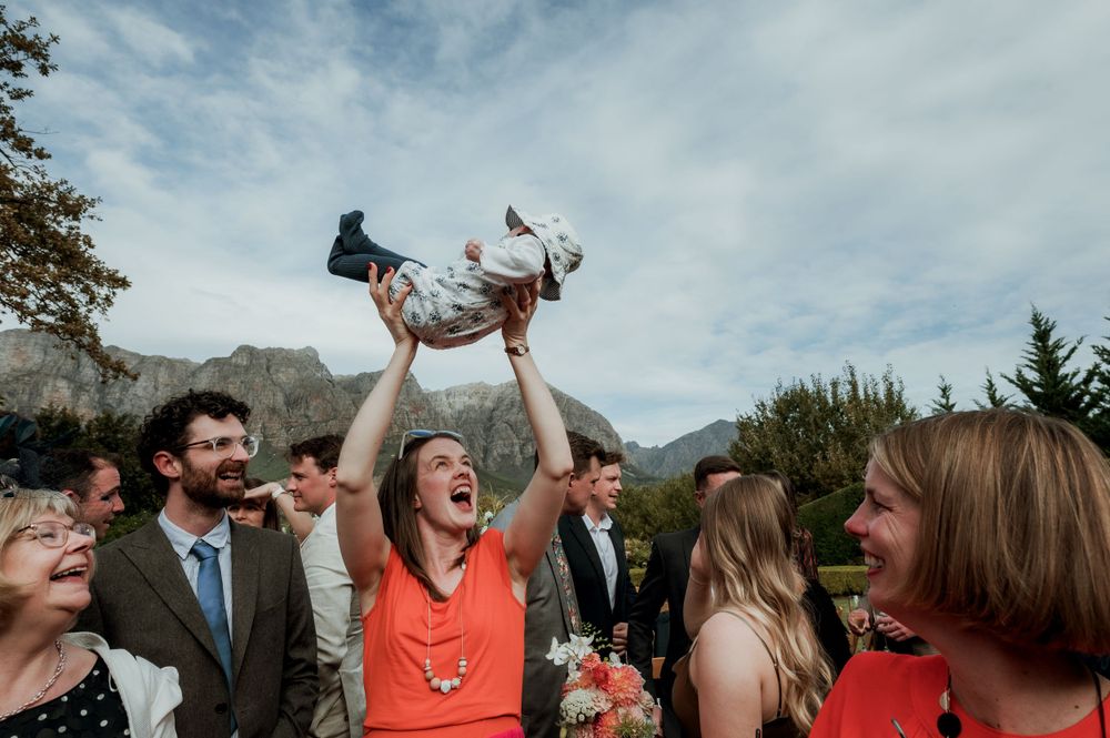 A woman in a bright orange dress lifts up and celebrates with a baby outdoors with mountains in the background at Johannesdal 1207