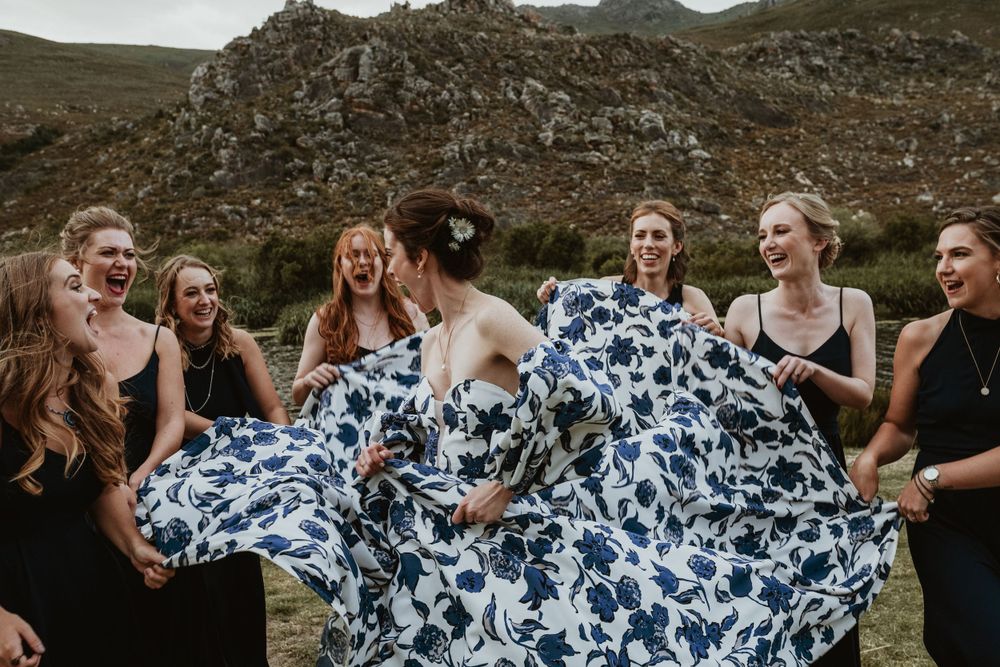 Wedding party in black dresses admiring bride's flowing blue and white patterned dress against mountain backdrop.
