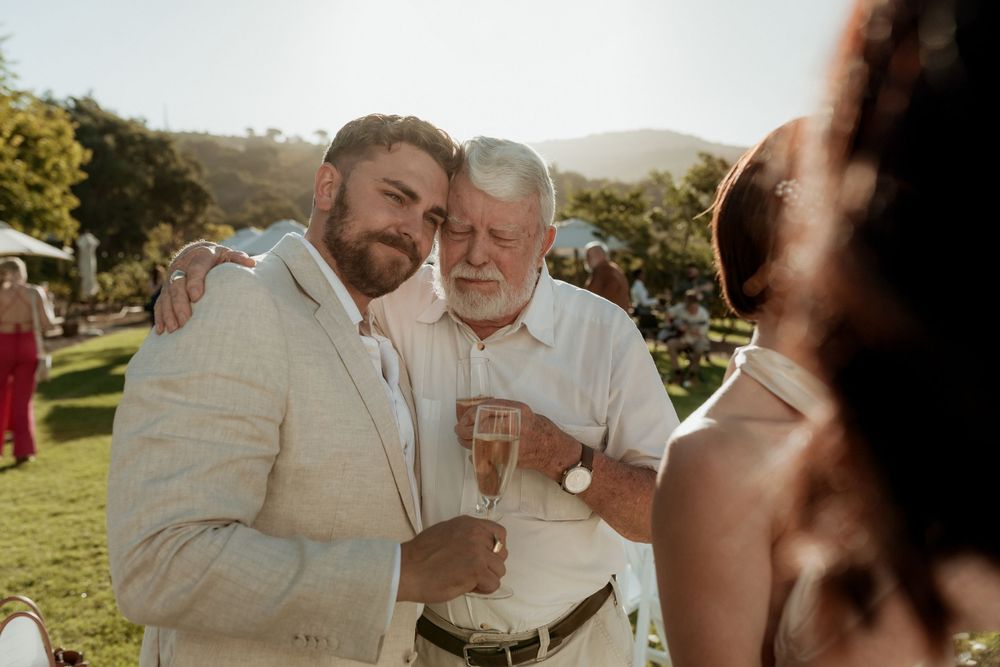 Two celebratory guests share a heartfelt moment while holding champagne glasses at an outdoor wedding reception.