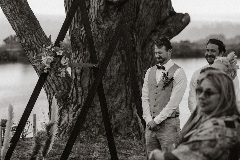 Black and white wedding ceremony photo taken outdoors by a large tree trunk near water. The groom is emotional and crying