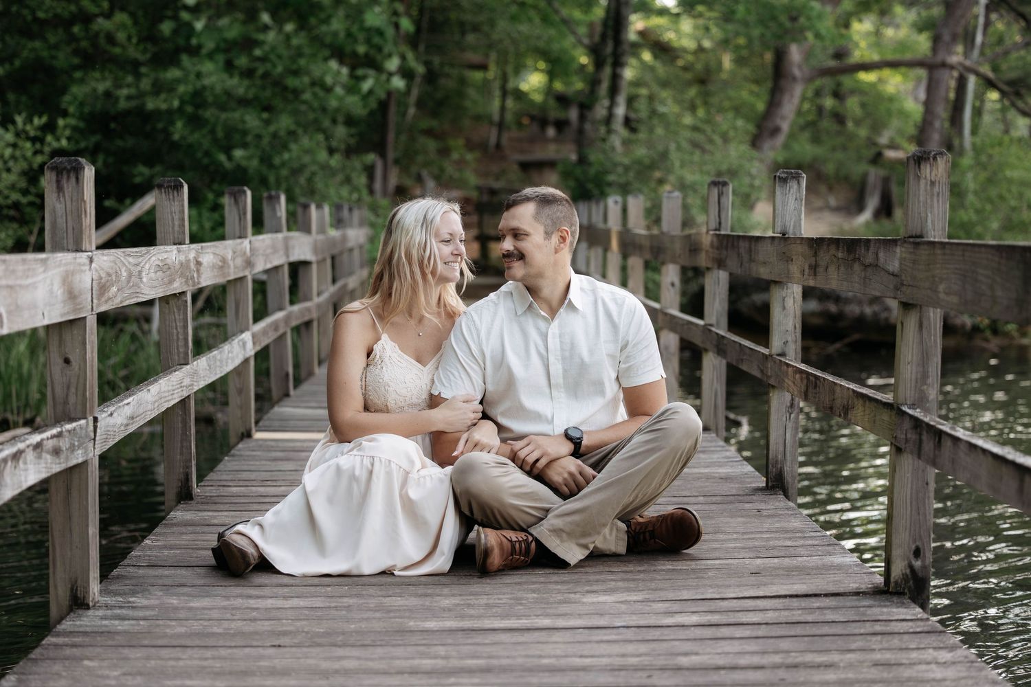 Couple sitting on wooden bridge dock, smiling at each other, surrounded by lush green trees near water.