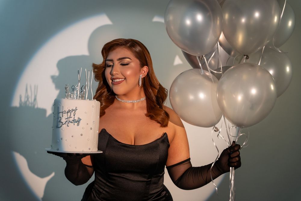 Elegant birthday portrait in studio holding a cake and balloons under spotlight lighting.