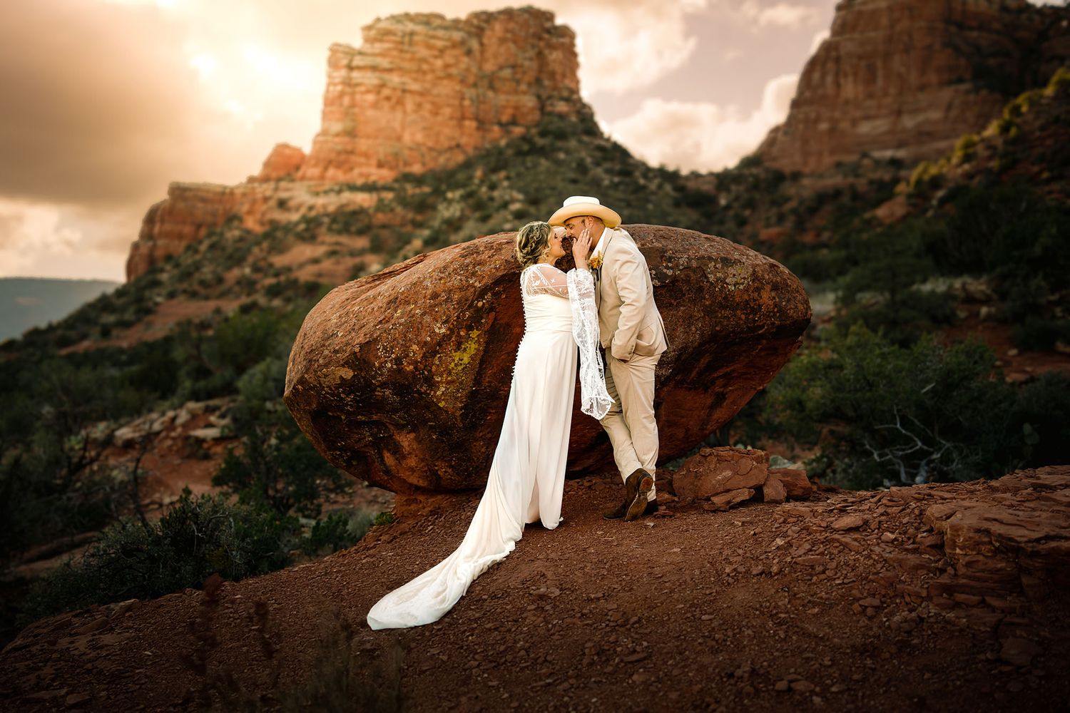 Couple sharing a romantic kiss at golden sunset in desert landscape, bride in white dress, groom in gray suit.