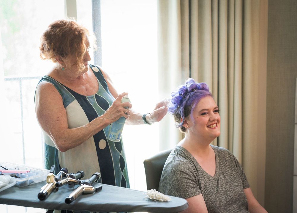 Hairstylist styling purple hair of smiling woman seated at vanity table during bridal preparation.
