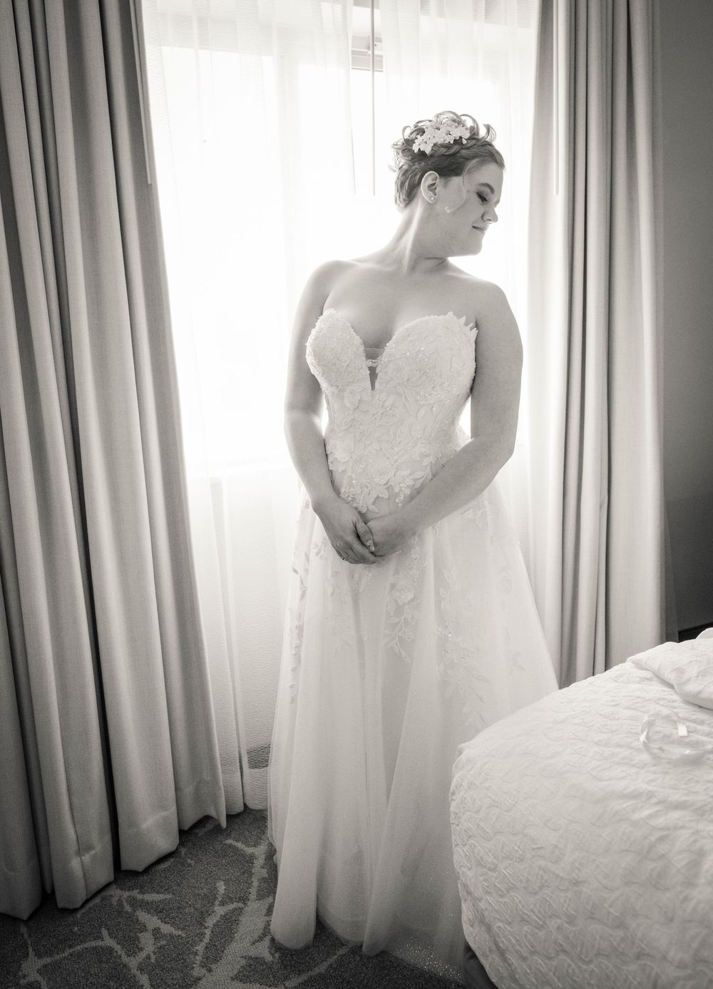 Bride in lace sweetheart wedding gown stands by window in black and white portrait photo.