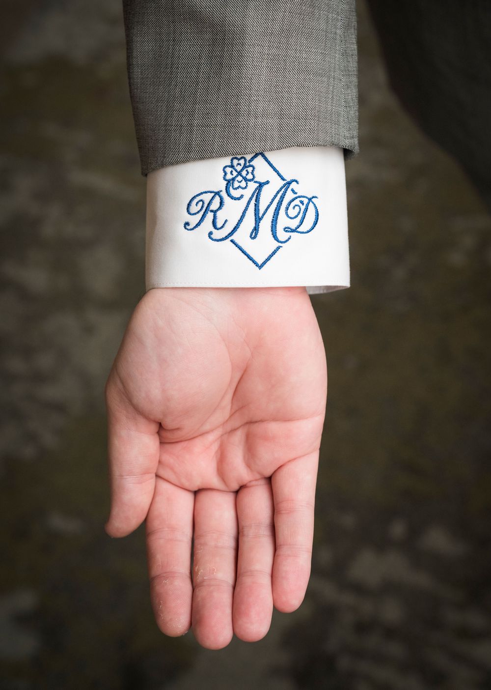 Groom's shirt cuff monogrammed with RMD in blue on white, sleeve of gray suit jacket visible.