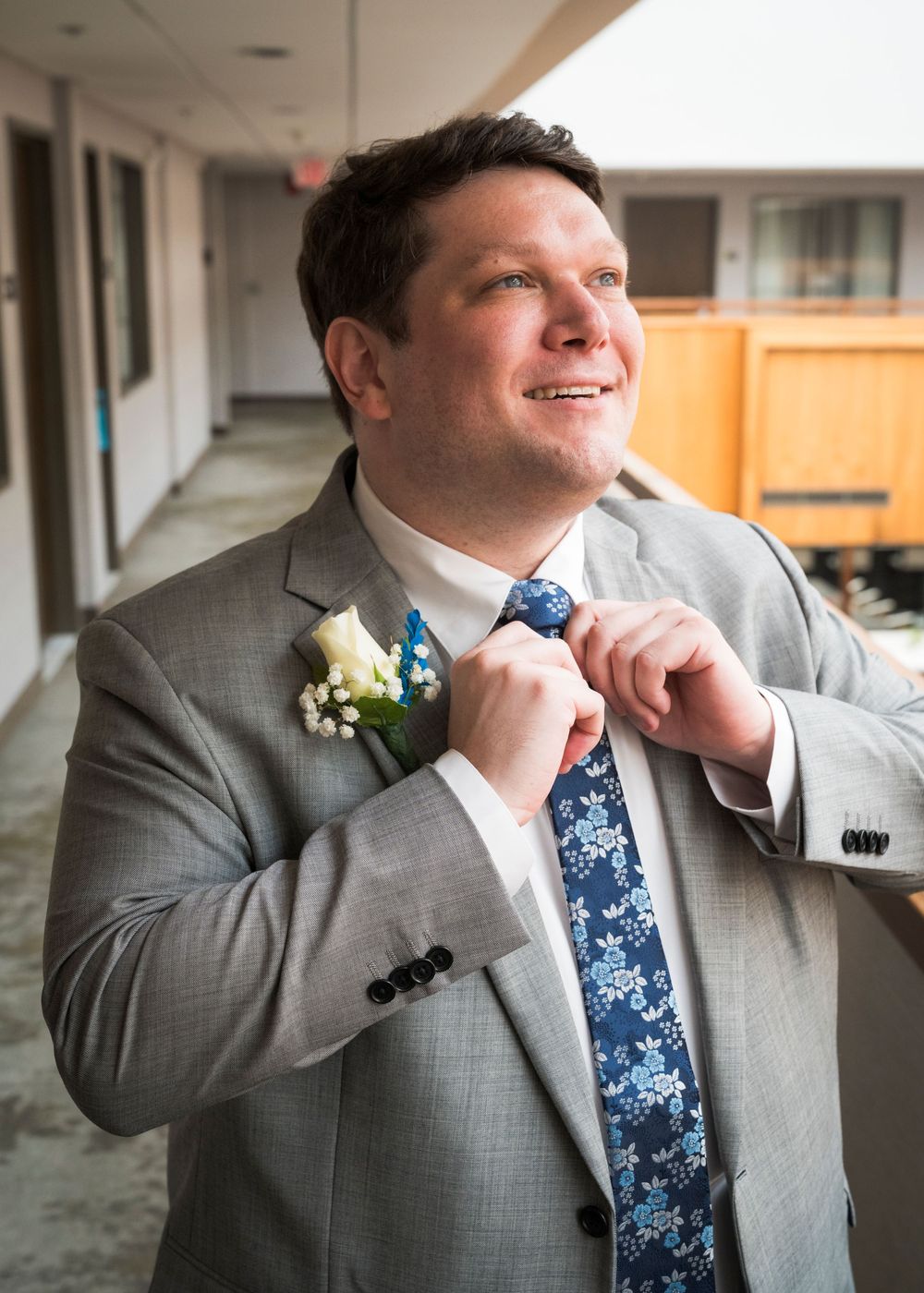 Smiling groom in gray suit adjusting floral tie with boutonniere in hotel corridor.