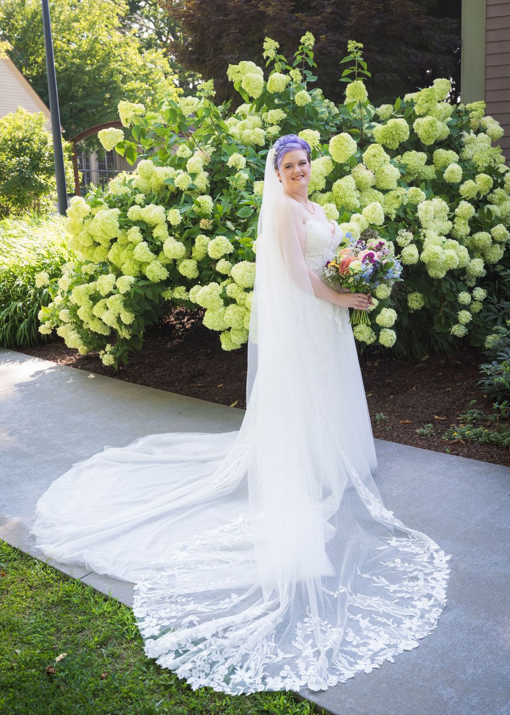 Bride in elegant white lace wedding gown with long cathedral veil holding bouquet in front of blooming hydrangeas.