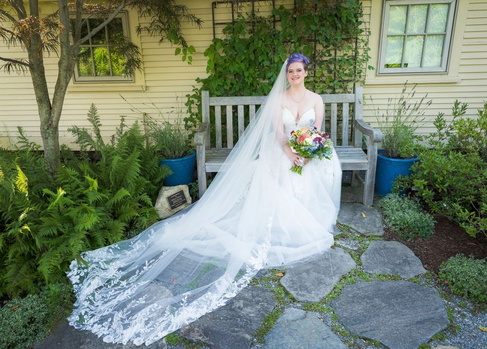 Bride in a stunning white gown with long lace veil holding a colorful bouquet in a garden setting.