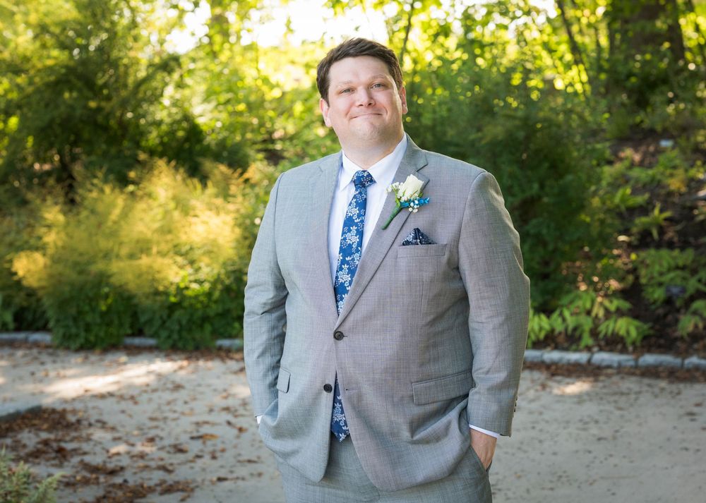 Man in gray suit with floral tie and boutonniere posing outdoors at a wedding ceremony.