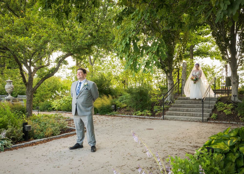 Groom in gray suit awaits bride descending garden stairs for romantic first look moment.