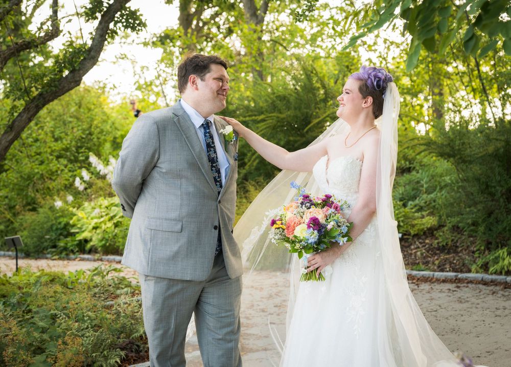 Bride with purple hair holding colorful bouquet touches groom's boutonniere in gray suit at outdoor wedding ceremony.