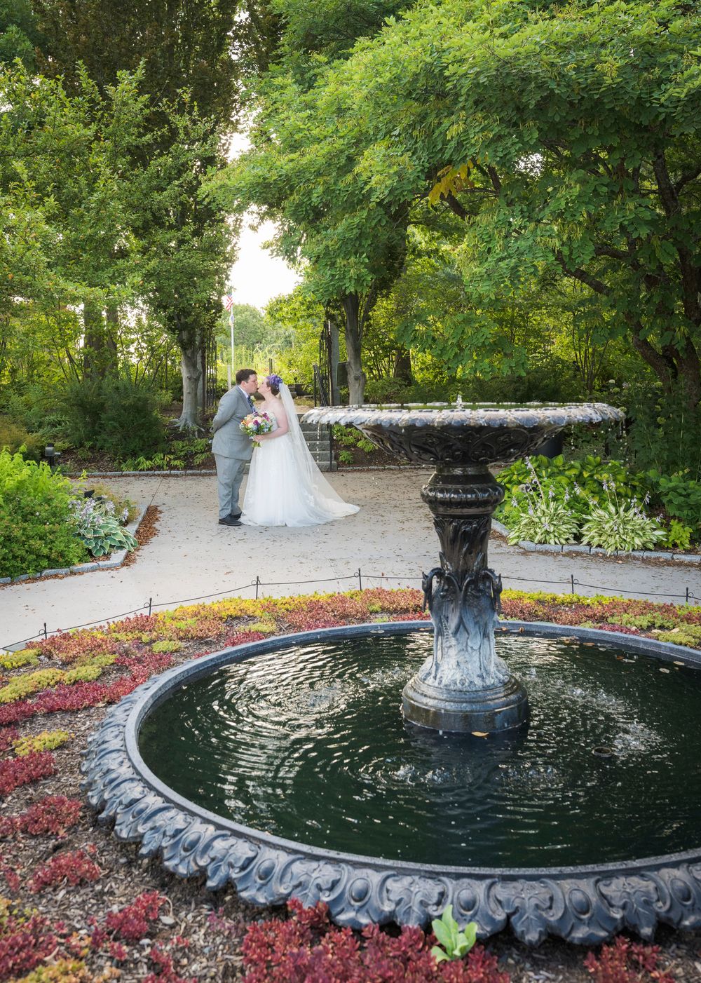 Bride and groom kissing near an ornate garden fountain surrounded by colorful flowers and lush green trees.