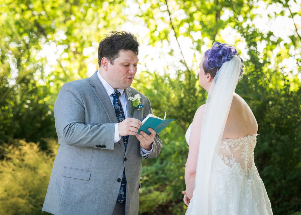 Groom in gray suit reads vows from teal book to bride in lace wedding dress with purple hair outdoors.