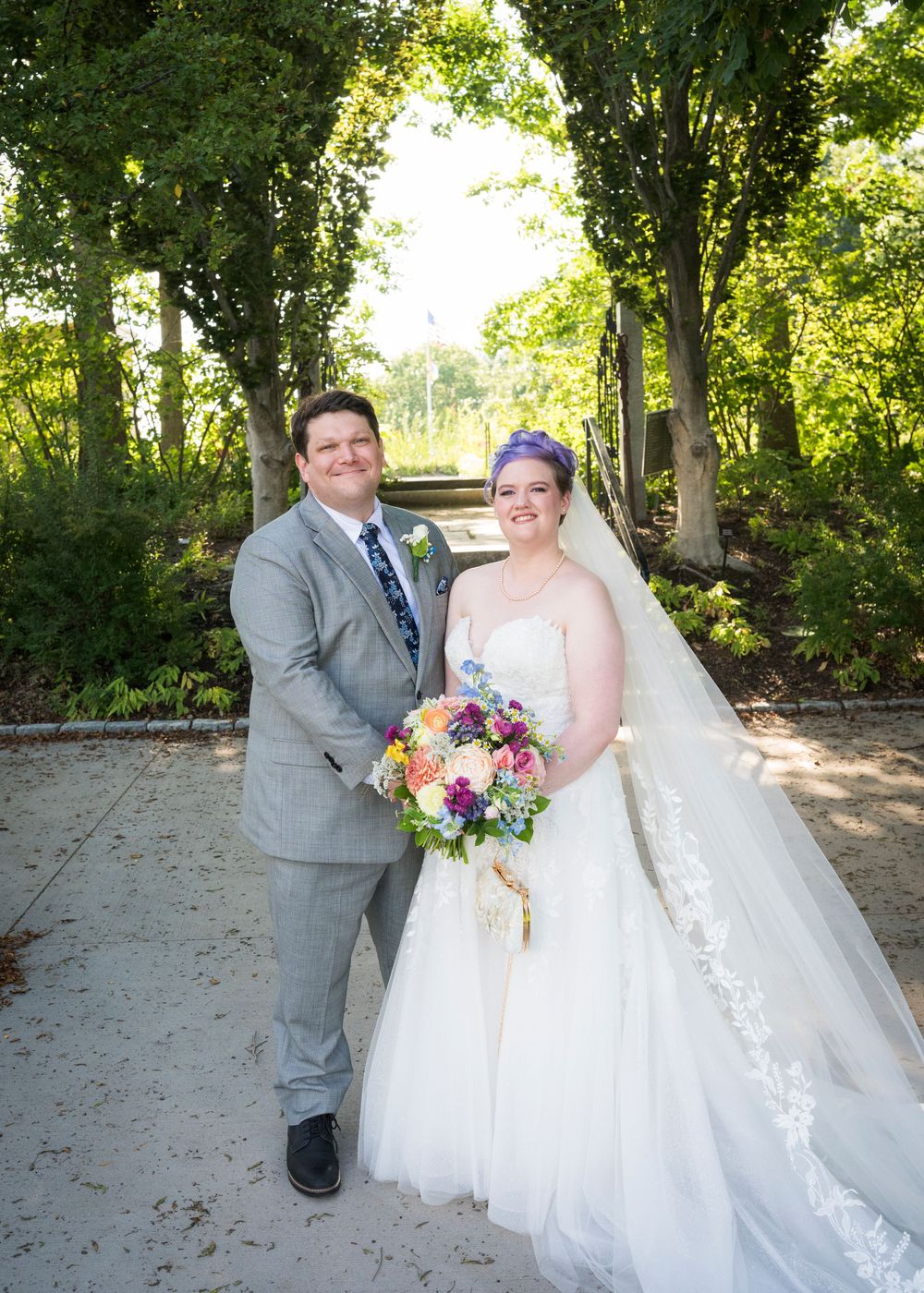 Bride in white gown with veil holds colorful bouquet beside groom in gray suit in garden setting.