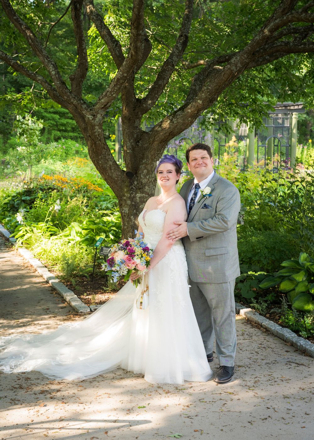 Bride and groom posing under a large tree in a lush garden, bride holding a colorful bouquet in a white gown.