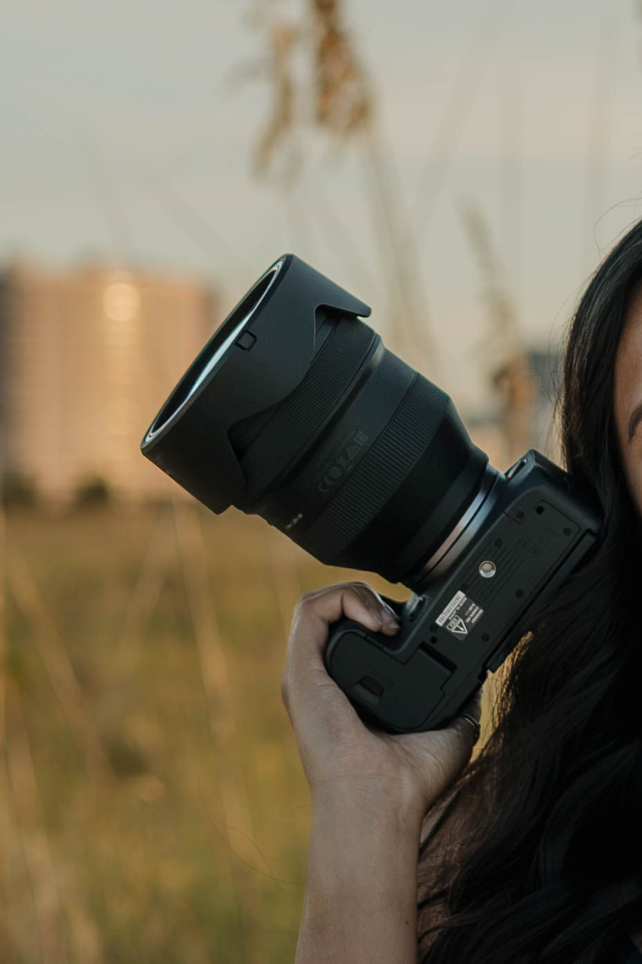 A woman with long dark hair holds a professional DSLR camera with a large lens outdoors in a golden field.