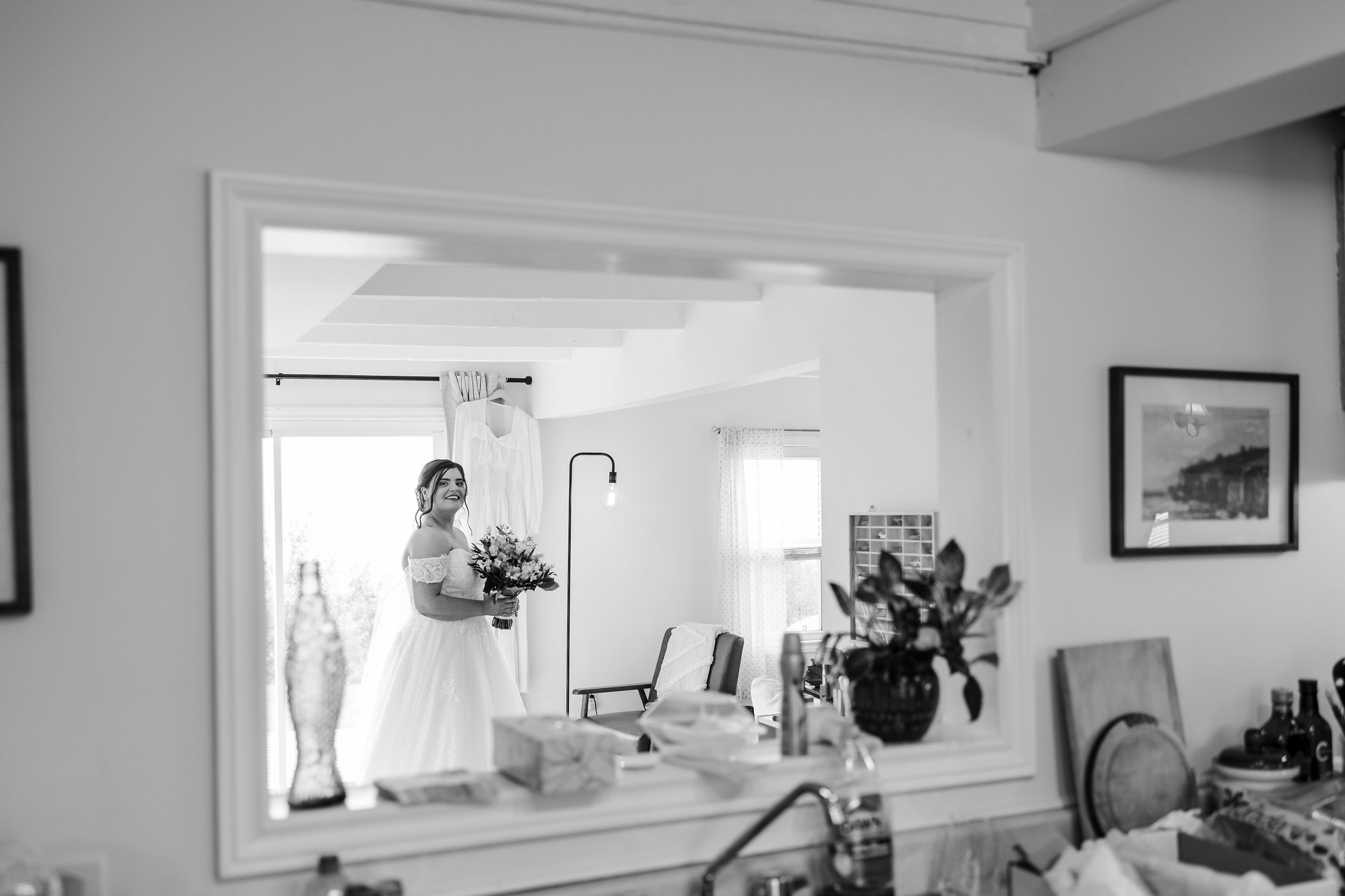 Black and white photo of a bride holding a bouquet reflected in a large mirror during wedding preparations.