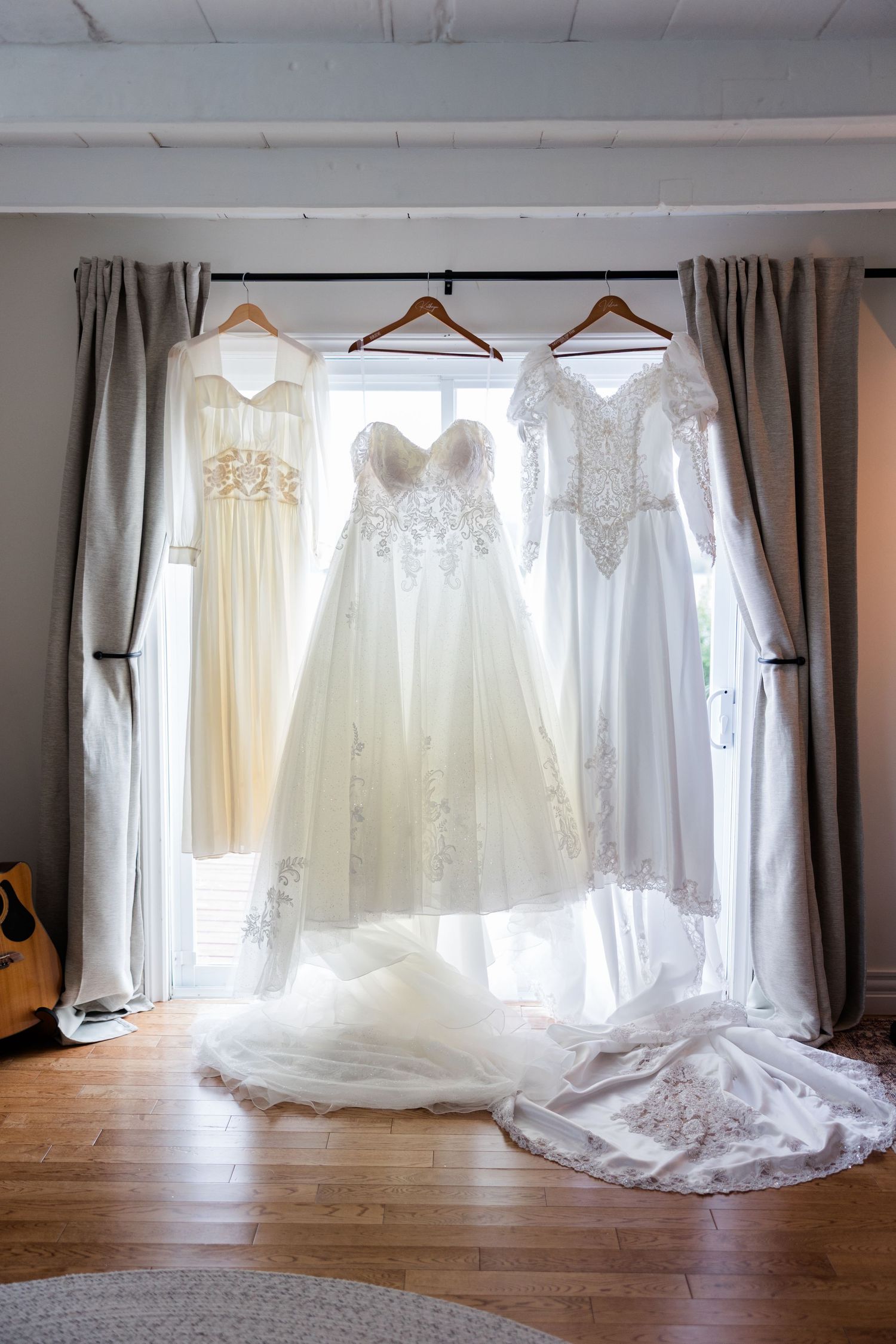 Three elegant white wedding gowns with lace details hanging on wooden hangers in front of a bright window.