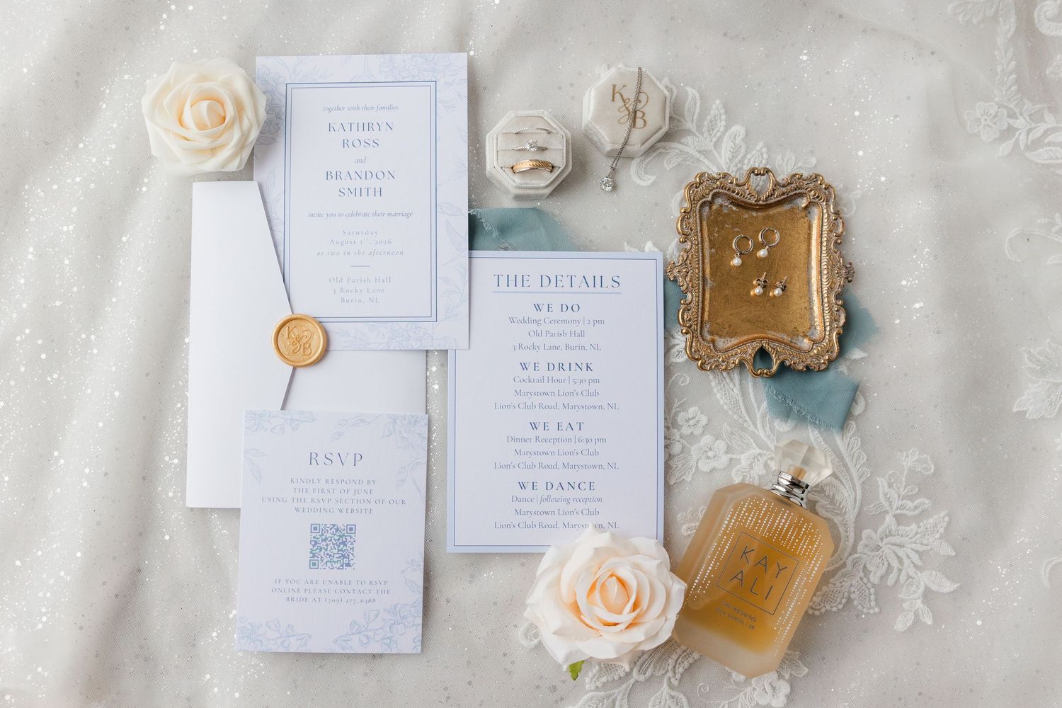 Elegant wedding stationery flatlay with invitations, RSVP card, wax seal, roses, perfume bottle, and gold jewelry tray.