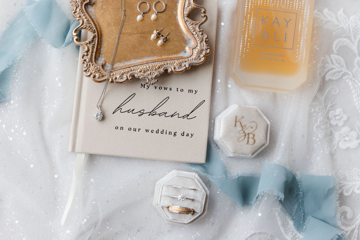 Wedding flat lay with vow book, gold rings in white velvet box, pearl earrings, ribbon, and perfume on blue fabric.
