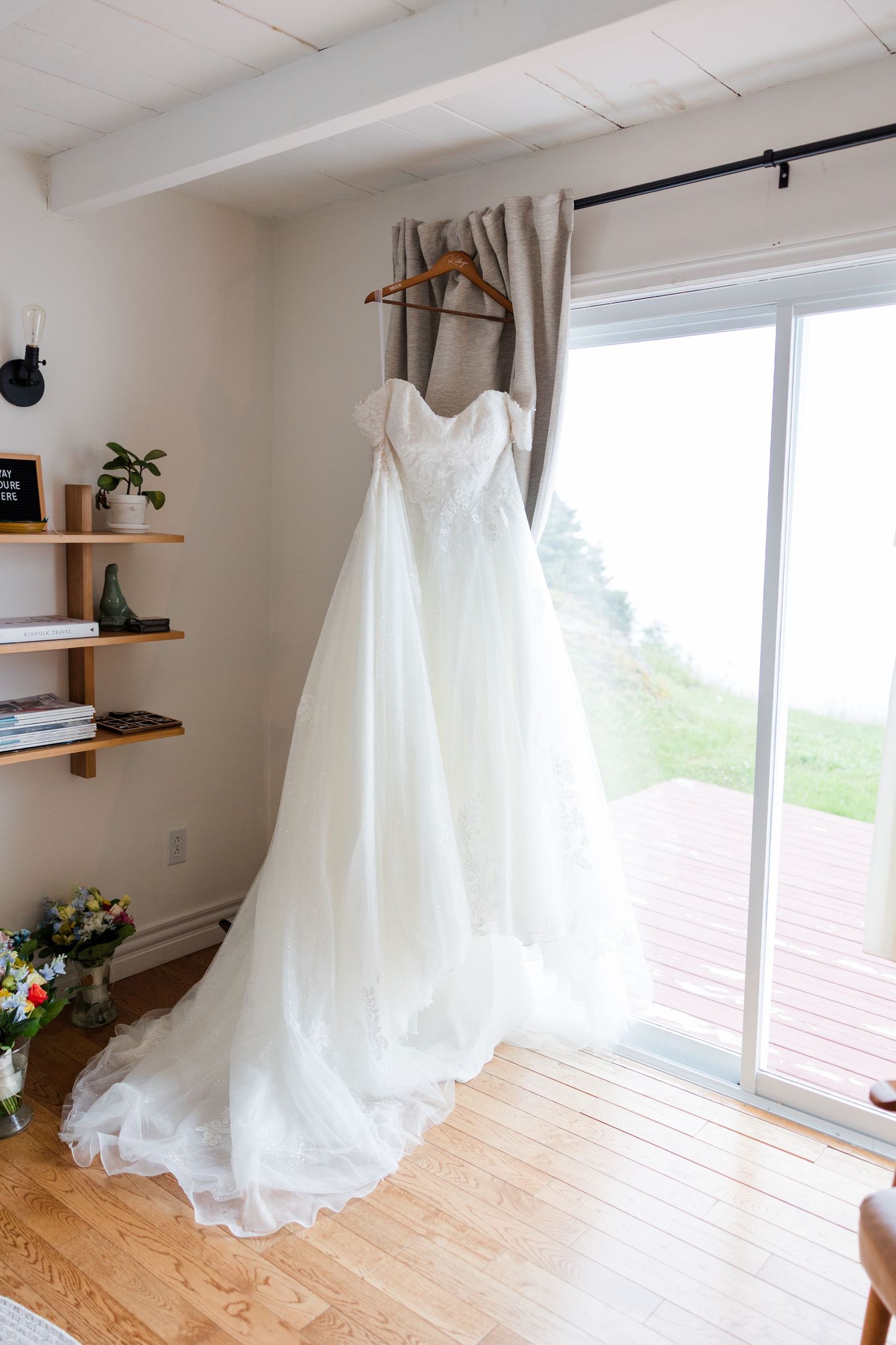 White wedding gown with lace bodice hanging on wooden hanger in bright room with hardwood floors and sliding glass door.
