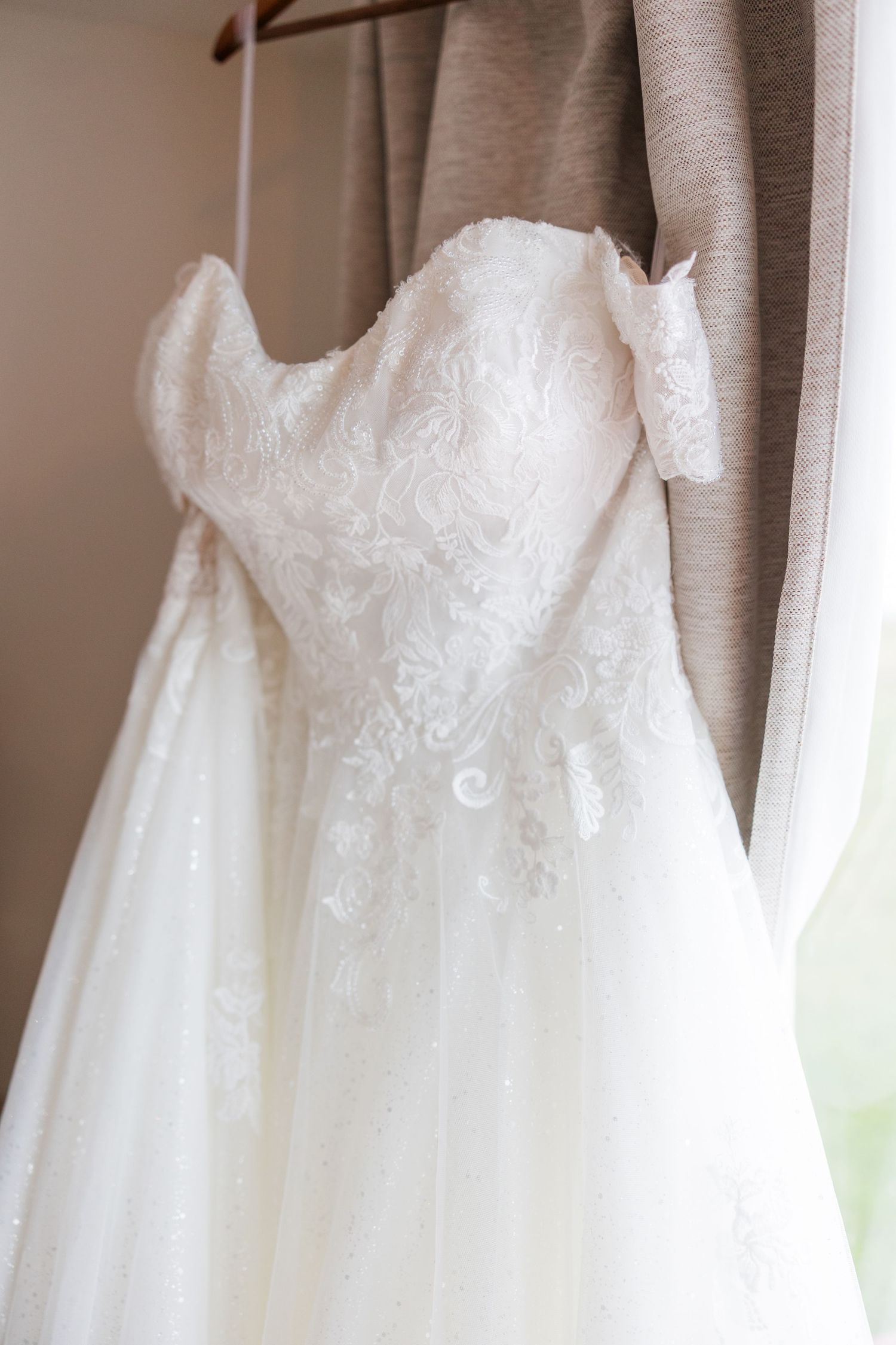 White lace strapless wedding dress hanging on a wooden hanger near a window with soft natural light.